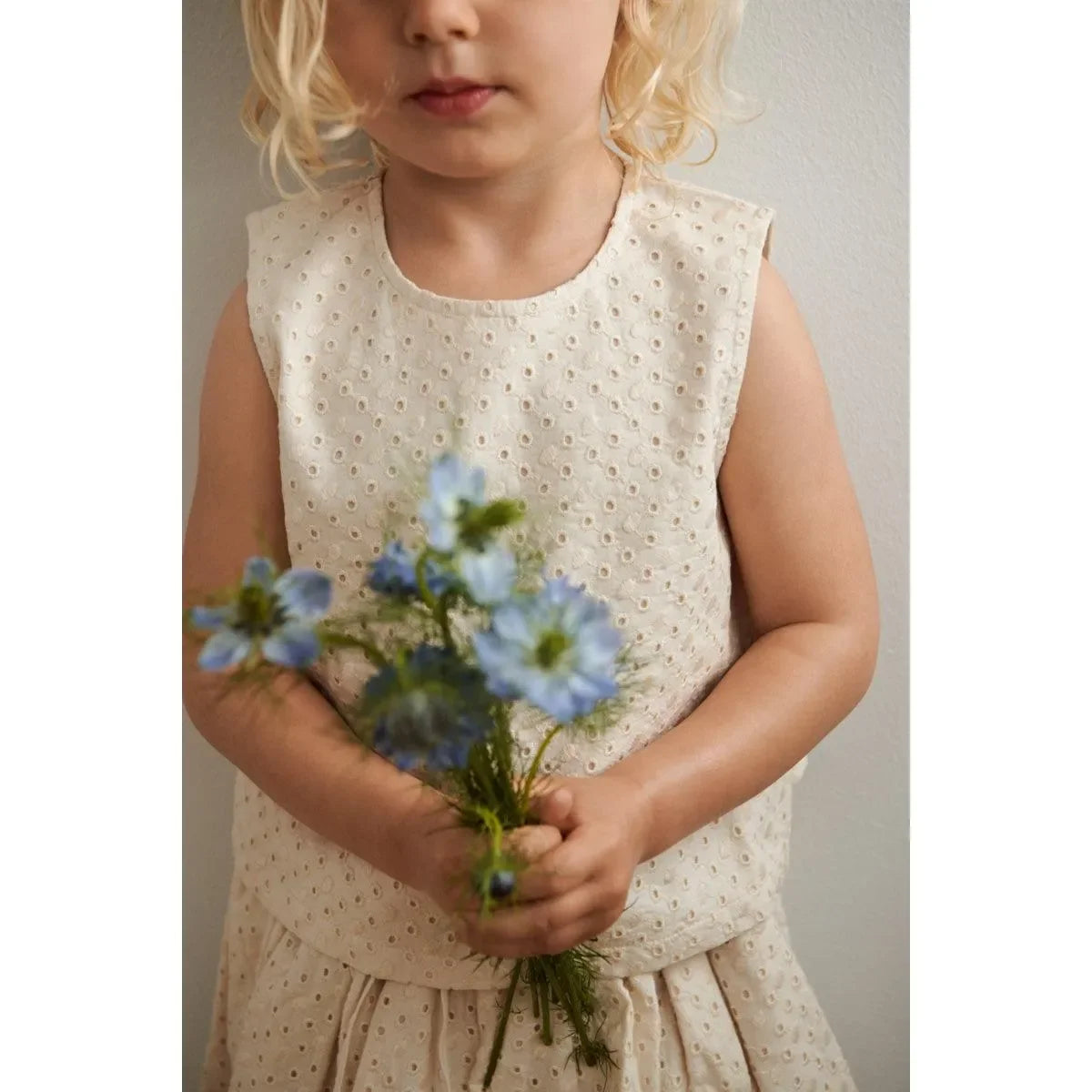 Kind im cremefarbenen Lochstickerei-Top hält blaue Blumen, minimalistische Wand im Hintergrund
