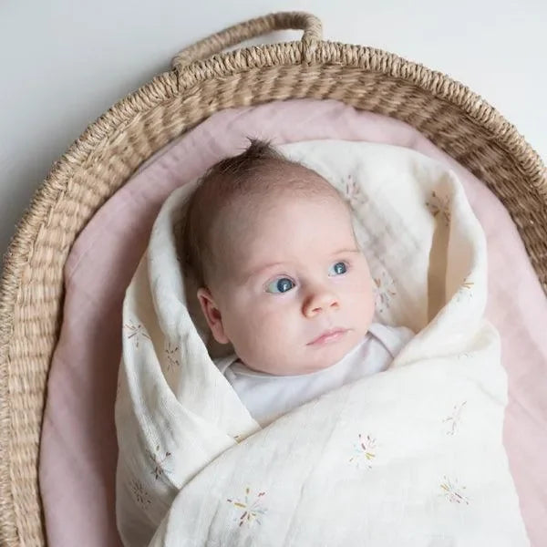 Baby in weicher Musselin-Babydecke mit Regenbogenmuster in Moseskorb auf rosa Unterlage
