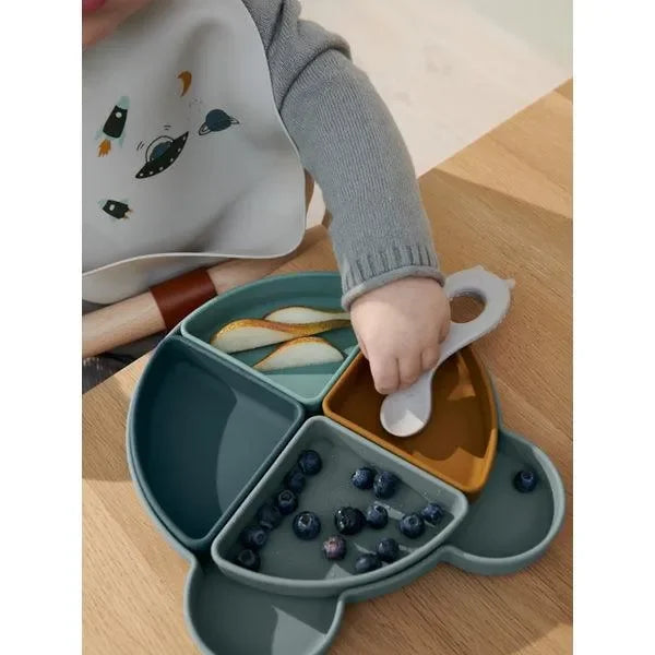 Baby greift mit Löffel nach Essen auf unterteilter Bärenteller aus Silikon, Blaubeeren und Birne