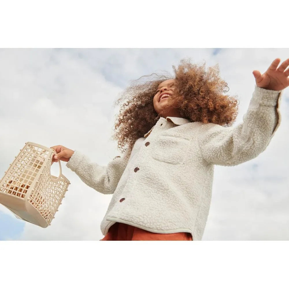 Kind mit lockigem Haar und cremefarbener Jacke hält beige Kinderkorb im Freien