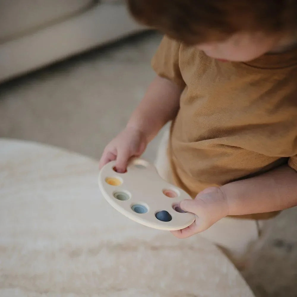 Kind hält Montessori Silikon Spielzeug in Form einer Farbpalette mit bunten Knöpfen