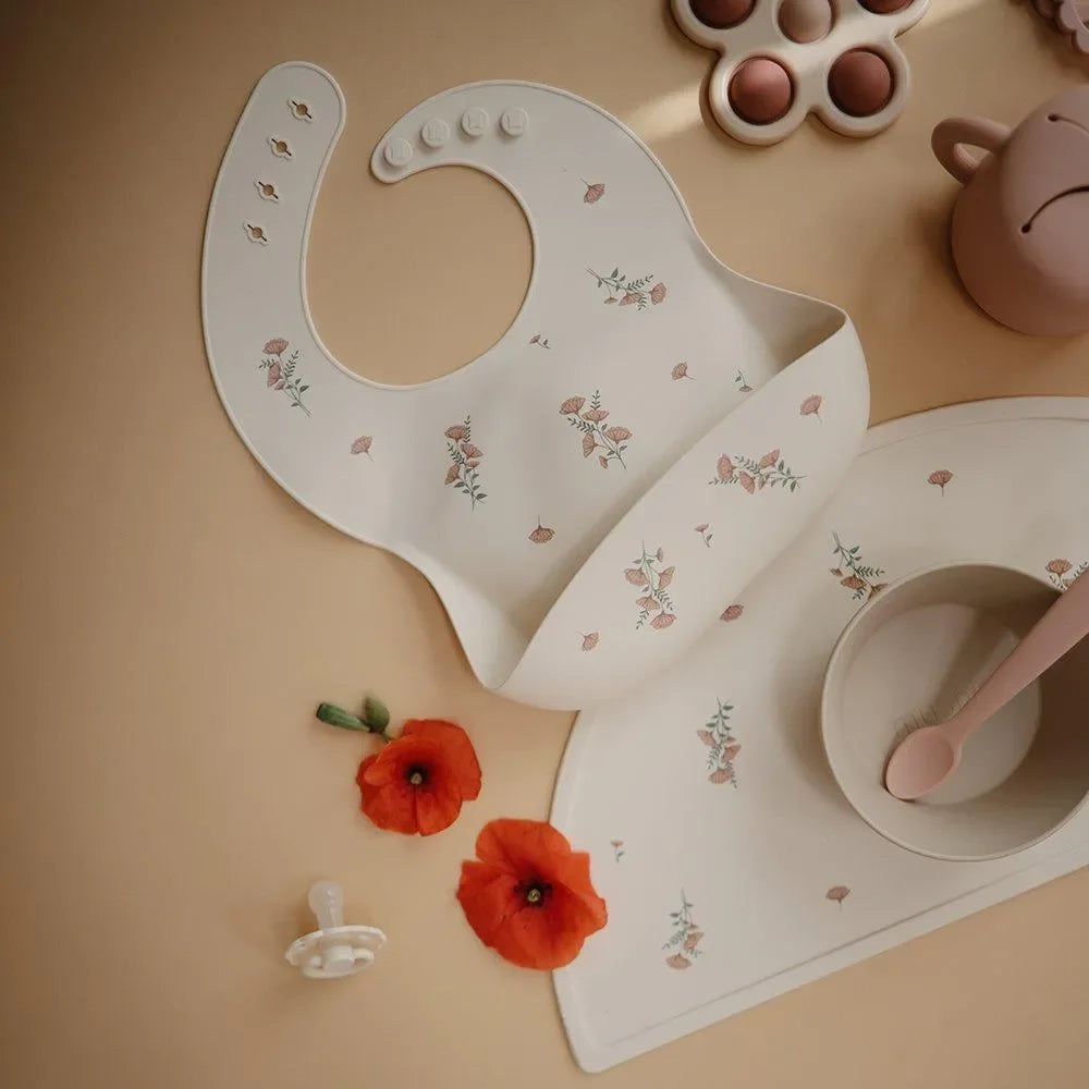 Beiges Silikonlätzchen mit rosa Blumenmuster, daneben Babygeschirr und Schnuller auf Tisch
