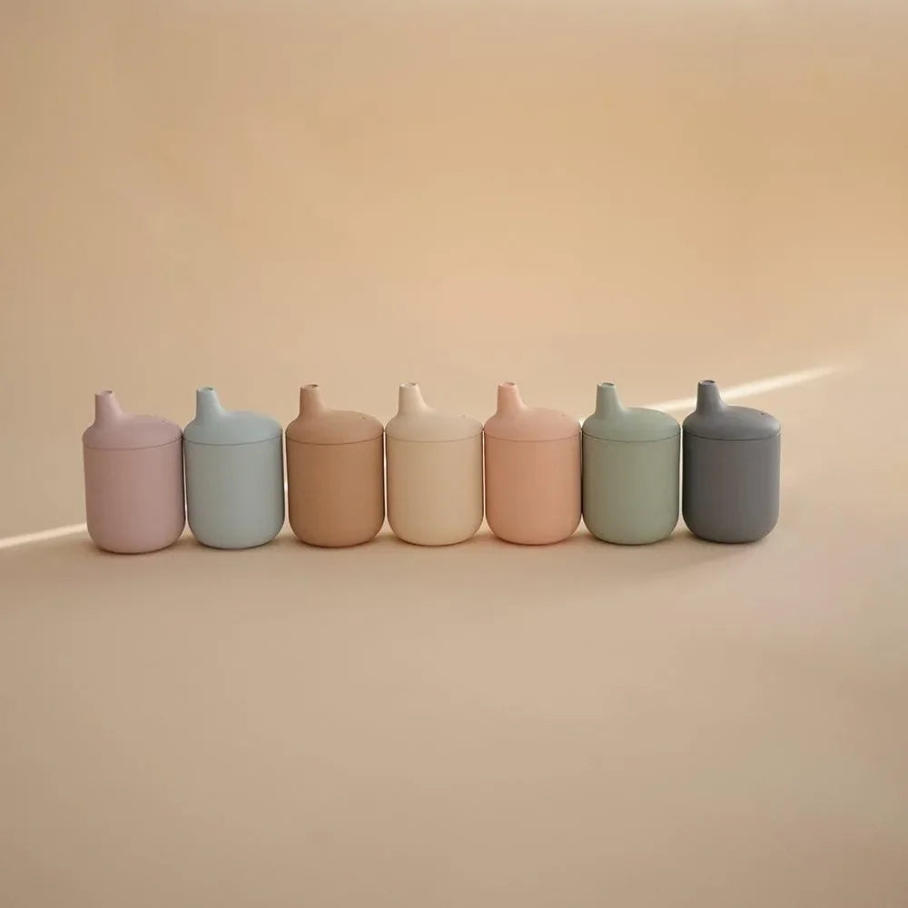 Sieben bunte Silikon-Trinkbecher für Kinder mit Deckel, auf beigem Hintergrund
