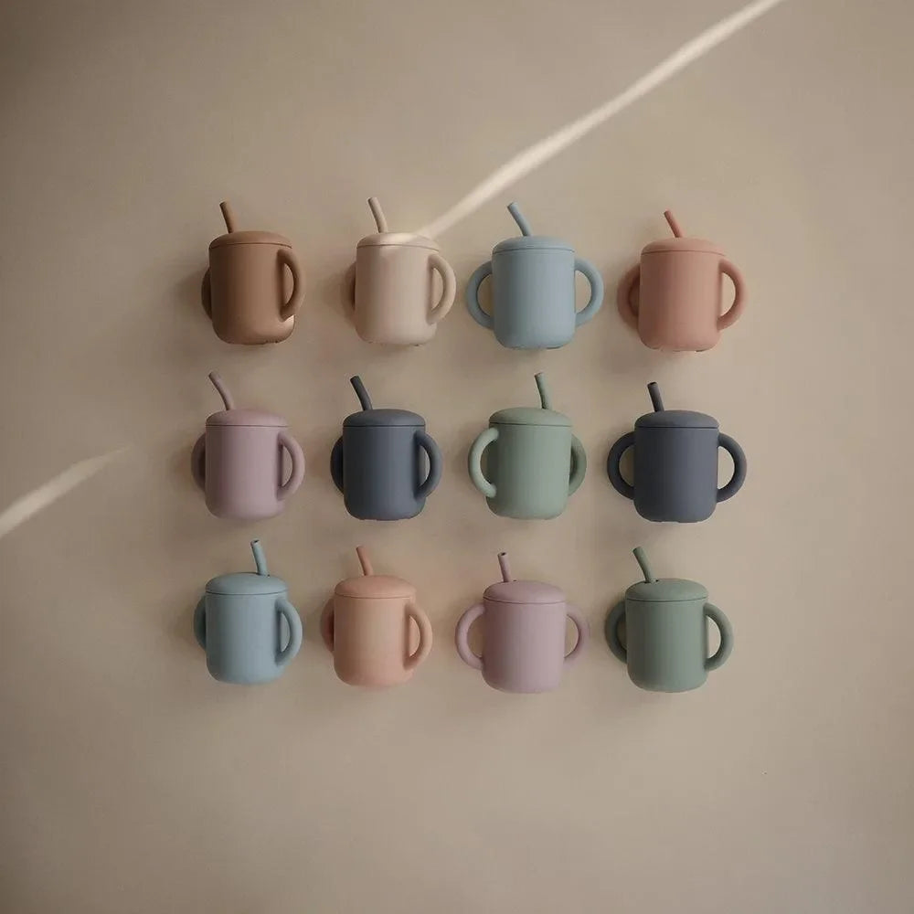 Zwölf bunte Silikon-Trinklernbecher mit Strohhalm und Griffen für Kinder vor neutralem Hintergrund