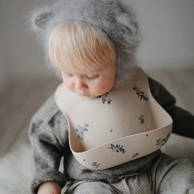 Baby mit beiger Lätzchen mit Blumenmuster und grauer Mütze, sitzt auf hellem Boden